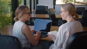 Two businesswomen are working together on a laptop in a modern office, with one typing and the other holding a notebook, surrounded by plants, creating a productive and natural workspace video