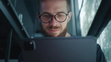 Young businessman with eyeglasses and beard smiling while using tablet in modern office, showcasing joy of success and excitement of entrepreneurial achievement. IT businessman video