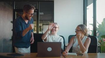 Three happy colleagues are giving high five to each other in office, celebrating their success after completing project using laptop. Concept Successful completion of transaction by sales team video