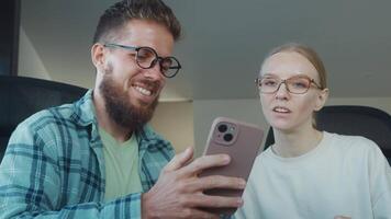 Two colleagues, man and woman, are sitting at their desks in office, sharing funny moment as they look at smartphone together, enjoying lighthearted break from work. Enjoyment of funny short videos