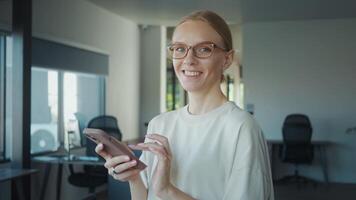 administrativo asistente vistiendo los anteojos utilizando móvil teléfono en moderno oficina, sonriente y hojeada Internet o social medios de comunicación, disfrutando descanso hora a trabajar. trabajo administrativo asistente video