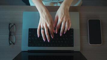 Womans hands typing on laptop keyboard on wooden desk, with smartphone and eyeglasses next to it, suggesting a modern work from home setup. Woman typing on laptop in home office, digital workflow. video