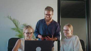 Tres alegre colegas son colaborando en proyecto, utilizando ordenador portátil y tableta, intercambiando ideas y trabajando juntos en contemporáneo oficina ambiente. diversidad y intervención de oficina equipo video