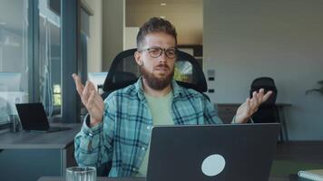 Young businessman shrugging and gesturing in confusion while working on laptop in modern office, expressing uncertainty and bewilderment. concept of negative emotions and misunderstandings in office video