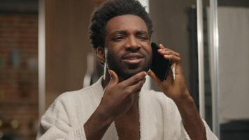 African American man looking at mirror reflection bathroom guy talking mobile phone touching face care shaving beard smiling bearded male call conversation smartphone communication in hotel bath home video