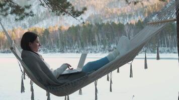Freelancer working remotely on laptop while relaxing in hammock, nestled beside frozen lake amid snow-covered pine trees, embodying digital nomad lifestyle and winter tranquility video