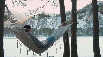 Young woman enjoys reading a book while relaxing in a hammock strung between two pine trees on the shore of a beautiful frozen lake in a tranquil winter landscape video