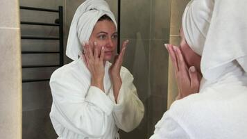 Beautiful woman wearing bathrobe and towel on head gently massages face skin with moisturizing cream, enjoying morning beauty routine in front of mirror in bathroom video