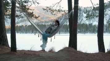 Comfortable woman wearing sweater, jeans and warm boots enjoys swinging in hammock strung between pine trees on shore of frozen lake surrounded by snowy winter forest video