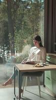 Young woman sitting at a table, enjoying breakfast and writing in a notebook by a large window with a view of a forest, illuminated by natural light video