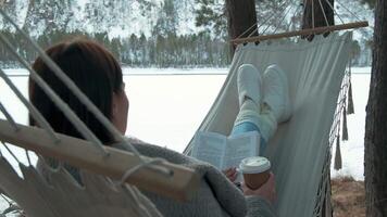 Young woman enjoys a peaceful moment, relaxing in a hammock by a frozen lake, immersed in a book and sipping coffee, surrounded by a serene winter landscape video