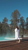Serene winter landscape featuring young woman removing bathrobe and entering steaming turquoise thermal pool surrounded by snow-covered pine forest under pristine blue sky video