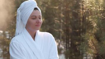 Wellness-focused woman wrapped in bathrobe and towel standing amid snowy winter forest, breathing deeply while creating visible vapor against serene, frost-covered landscape video
