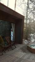 Cozy morning ritual features a woman in a bathrobe, savoring a warm drink on the deck of a stylish, modern cabin nestled in a snowy woodland setting video