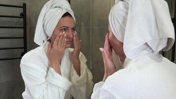 Young woman wearing white bathrobe and towel wrapped on head, gently applying moisturizing cream while looking in bathroom mirror during morning skincare routine video
