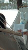 Cozy winter scene of a woman enjoying a peaceful moment in a hammock by a frozen lake, reading a book and sipping coffee, surrounded by snow-covered trees video