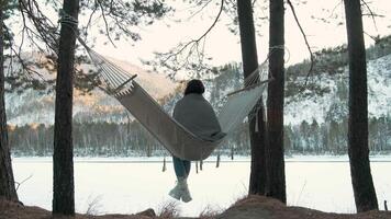 Winter landscape featuring young woman relaxing in hammock strung between pine trees, overlooking frozen lake during serene sunset with snow-covered surroundings video