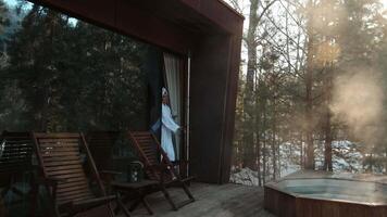Sunlight streams through the trees, illuminating the steam rising from a hot tub as a woman in a white bathrobe sips from a cup, enjoying the serene winter landscape video