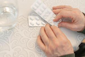 Senior person hands holding blister packs of pills over a patterned tablecloth, with a glass of water nearby, copy space. Concept of senior health care and daily medication routine photo