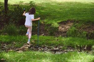Young Girl Crossing a Stream in a Green Forest Landscape Using Hiking Poles photo