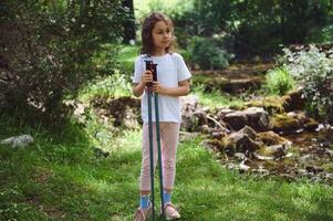 Young Girl Hiking in Forest Setting with Trekking Poles near Stream in Tranquil Nature Scene photo