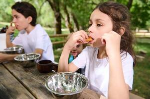 Children Enjoying Snacks Outdoors Around a Wooden Picnic Table in a Sunlit Forested Park Area photo