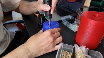 A Caucasian male in his 30s uses a power drill on a plastic container, surrounded by electronic components and a laptop DIY electronics and maker culture video
