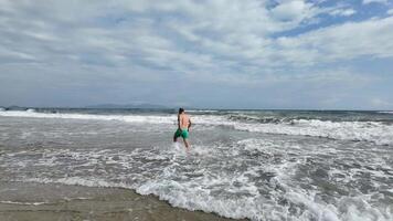 A Caucasian man in green shorts walks into the ocean on a cloudy summer day concepts include vacation and relaxation video