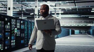 Data center system administrator tasked with reviewing code for AI automation scripts. African american man walking in server hub running diagnostics on equipment rigs, camera A video