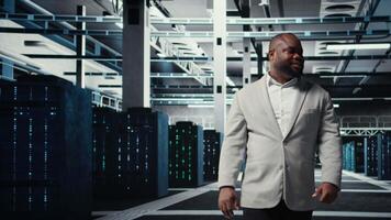 leende chef i data Centrum håller på med kolla upp på hårdvara till undvika driftstopp. Lycklig afrikansk amerikan handledare övervaka server rum infrastruktur superdatorer, kamera en video