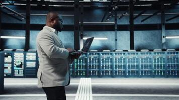Software developer in data center using artificial intelligence tech on laptop to deploy new server architectures. IT expert in server hub updating storage arrays using AI technology, camera A video