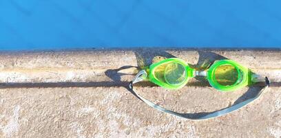 Bright green swimming goggles rest on the edge of a pool, casting a smile-shaped shadow in the sunlight. Perfect summer vibe and vacation mood photo