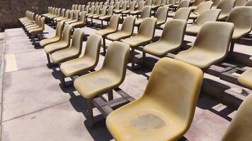 Rows of old, sun-faded plastic stadium seats in an outdoor arena under bright sunlight. The scene evokes a sense of summer heat and empty anticipation before an event photo