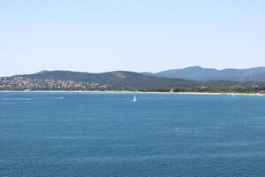 A peaceful coastal scene with a small sailboat floating on calm blue water. The distant shoreline is lined with green hills and scattered houses under a clear sky. photo