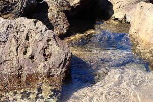 The image shows coastal rocks partially submerged in crystal-clear water. Sunlight reflects off the rippling surface, revealing the seabed and marine textures below. photo