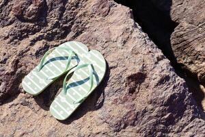 A pair of green flip-flops with white daisy patterns resting on a rough textured rock near the beach. Bright sunlight casts shadows, evoking a summer vacation mood. photo