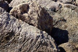 Textured coastal rocks with deep grooves and layers, shaped by centuries of wind and water erosion. The rugged surface contrasts with the soft shadows and sunlight. photo