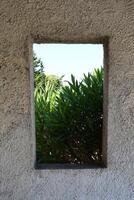 A view of vibrant green shrubs and tall trees framed by a rectangular window in a textured stone wall, capturing a peaceful moment of Mediterranean nature. photo