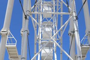 Low angle view of a white Ferris wheel structure with geometric metal supports and cabins, set against a vivid blue sky. The image captures architectural symmetry and summer leisure. photo