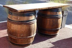 Two rustic wooden barrels with black metal bands support a thick wooden tabletop, forming a unique outdoor table setup on a red pavement, blending function and vintage style. photo