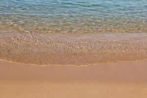 A gentle wave rolls onto a sandy beach under clear skies. The water is crystal-clear with shades of turquoise and blue, creating a peaceful and relaxing summer scene. photo