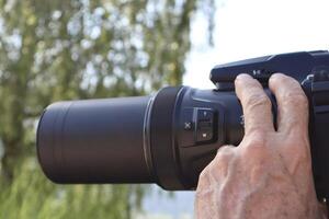 Detailed view of a male hand holding a professional recording device with a large zoom lens outdoors during a daytime shoot, focused on modern camera equipment photo