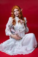 Elf cosplay young woman with long red hair sits on red background with white wicker basket on her lap. There is funny Sphinx kitten in basket. Elf in white summer dress looks at camera and smiles. photo