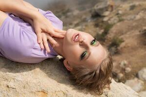 Splendid female fashion model lies on back on rock in mountains, places both palms on her neck and looks into camera. Magnificent sensuality Caucasian ethnicity woman in relaxed outdoor playful pose photo