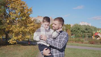 In a bright autumn park, a father gently holds his son, surrounded by colorful foliage and a playground, enjoying quality time on a sunny day. photo