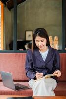 A young woman in a casual setting writes in a notebook with a laptop beside her, capturing a moment of focus and creativity. photo