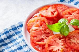 Tomato Slices with Basil Leaves on White Plate, Blue Checkered Napkin, Light Grey Background, Top View photo