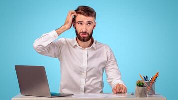 Confused millennial guy with word cloud above his head sitting at desk with laptop, blue studio background, mockup for design. Puzzled office worker with speech bubble, copy space photo