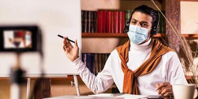 Distant Online Education. Indian Teacher Guy Making Lesson For Educational Blog Teaching English Grammar Rules, Pointing At Blackboard Sitting At Home, Wearing Face Mask. Selective Focus photo