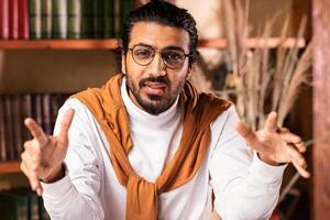 Arab Teacher Man Talking To Camera Teaching Online Explaining Lecture To Remote Students Sitting In Living Room Near Books Shelf Indoor, Working From Home. E-Teaching, Remote Tutoring. Selective Focus photo